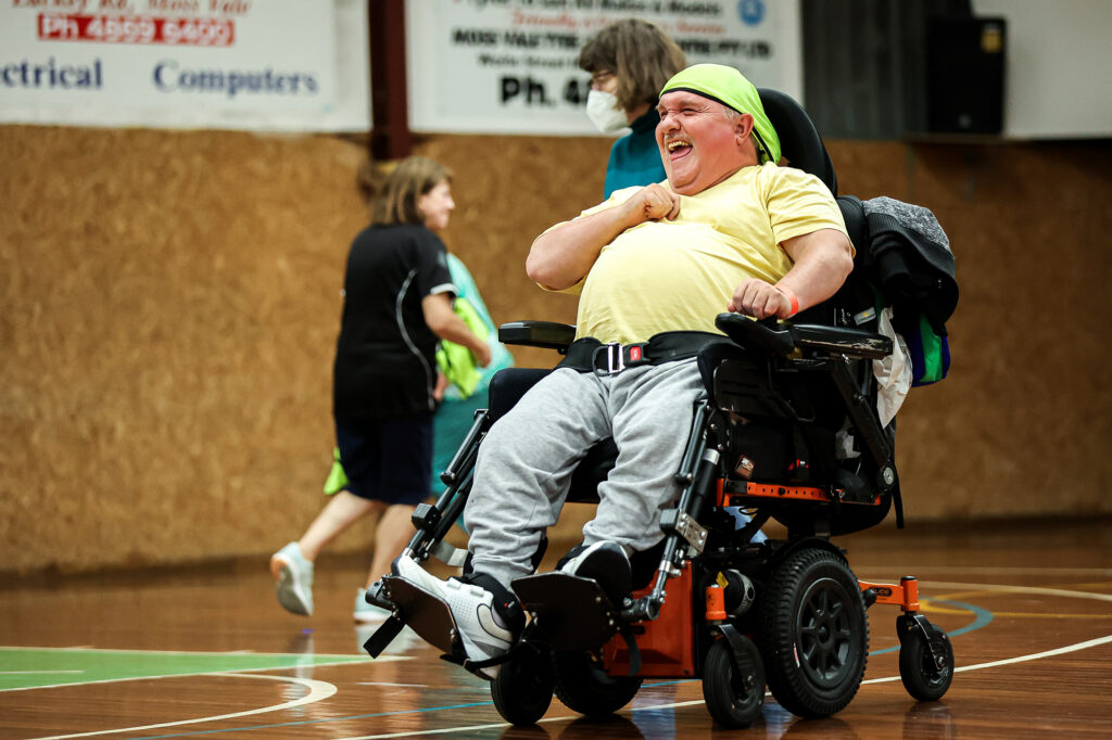 A man in a power wheelchair laughs joyfully while participating in an indoor sports activity. He wears a yellow shirt, grey sweatpants, and a green bandana. Other players and facilitators are in the background on a indoor court.